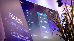 AKOS 2|5-Schild und "Tag 2"-Agenda bei einer Konferenz, mit einer Pflanze davor und blau-violetter Beleuchtung im Hintergrund.