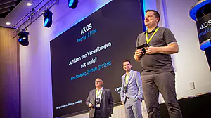 Drei Männer mit Namensschildern stehen auf einer Konferenzbühne, hinter der ein beleuchteter Bildschirm mit deutschen AKOS-Texten und Themen zu sehen ist.