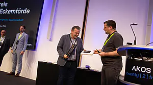 Bei einer Preisverleihung tauschen zwei Männer auf der Bühne eine Plakette aus, während im Hintergrund andere und Präsentationsfolien erscheinen.