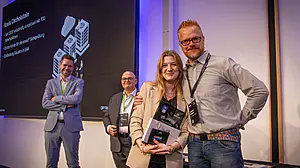 Vier Personen bei einer Indoor-Veranstaltung, eine Frau mit einer Plakette wird beglückwünscht. Zwei Männer stehen dahinter; großer Bildschirm mit Text sichtbar.