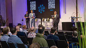 Das Publikum sieht drei Männer auf der Bühne, von denen einer spricht. QR-Codes und Social-Media-Symbole werden auf Bildschirmen in einem modernen Konferenzraum angezeigt.