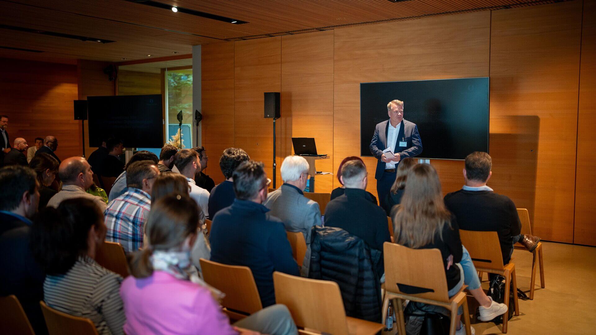 Ein Mann im Anzug präsentiert vor einem aufmerksamen Publikum in einem holzgetäfelten Konferenzraum, hinter ihm stehen ein Bildschirm und ein Laptop.