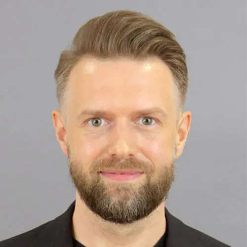 Smiling man with short light brown hair and a beard wears a black jacket, standing against a plain gray background.