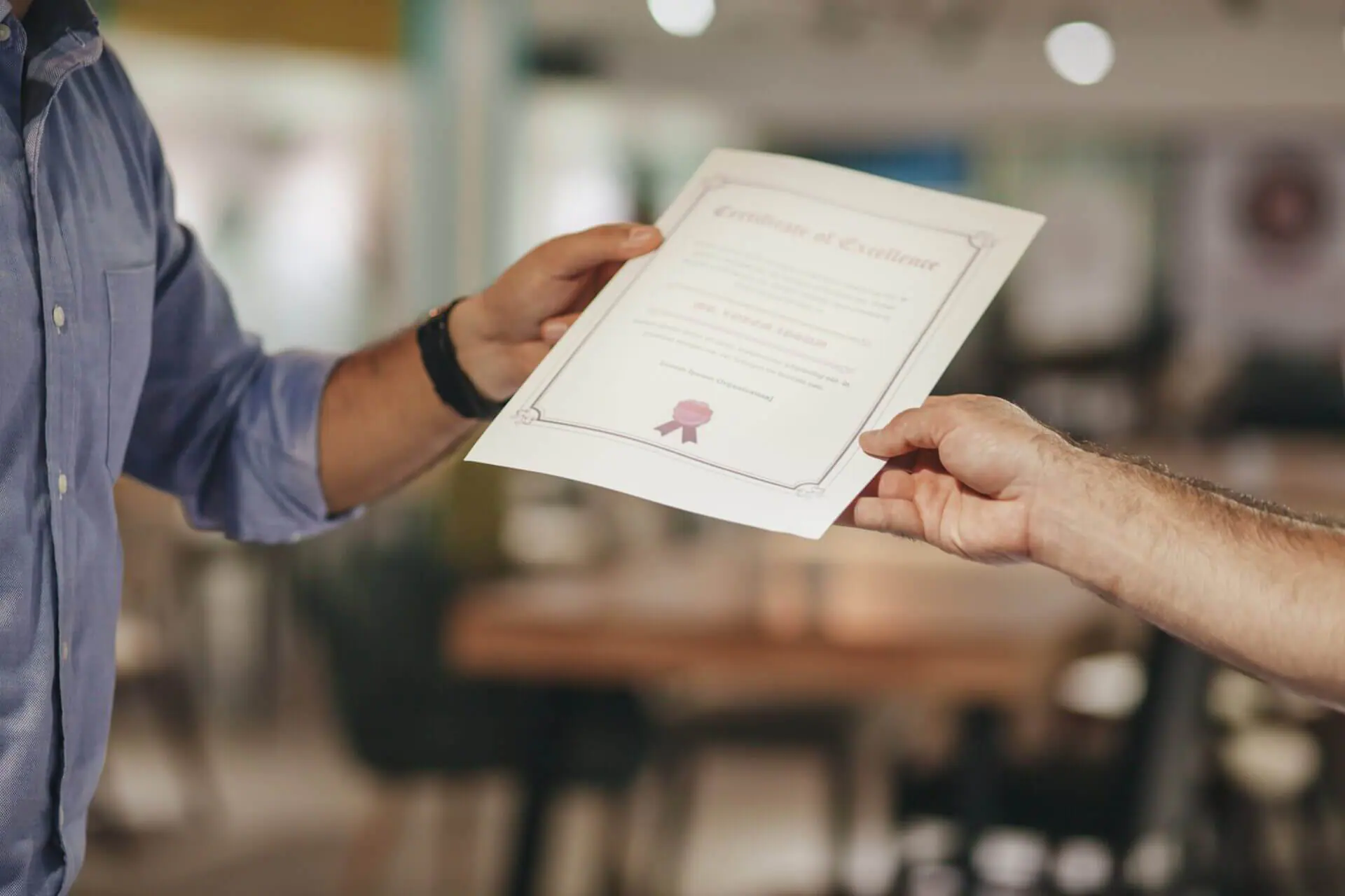 A person hands a certificate of excellence to another, both visible from the shoulders down in a blurred indoor setting.