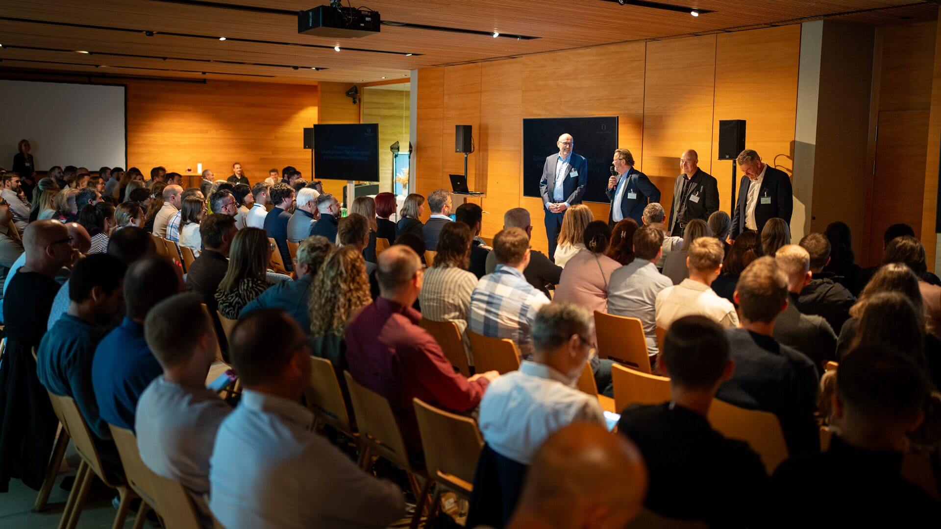 In einem holzgetäfelten Konferenzraum mit Stuhlreihen und Präsentationsgeräten steht das Publikum vor einer Bühne mit fünf Rednern.