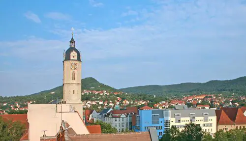 Uhrenturm mit dunkler Kuppel inmitten roter Dächer; grüne Hügel und ein teilweise bewölkter blauer Himmel im Hintergrund.