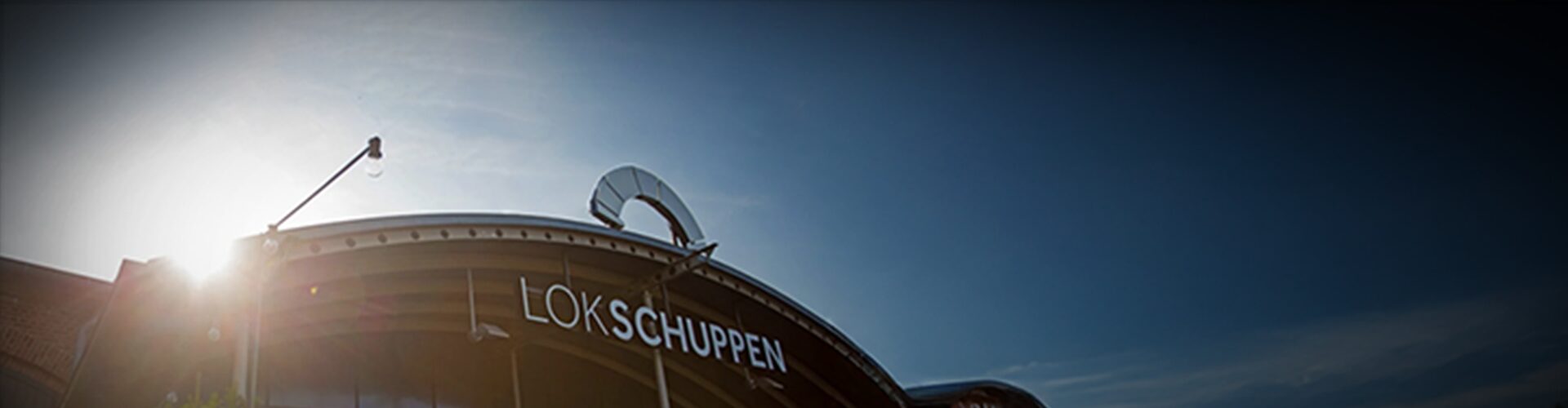Geschwungene Dachlinie mit "LOK SCHUPPEN" in fetten weißen Buchstaben unter einem klaren blauen Himmel; links leuchtet das Sonnenlicht.