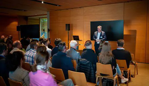 Ein Geschäftsmann hält eine Präsentation vor einer sitzenden Gruppe in einem modernen Konferenzraum, hinter ihm sind ein Bildschirm und ein Laptop zu sehen.