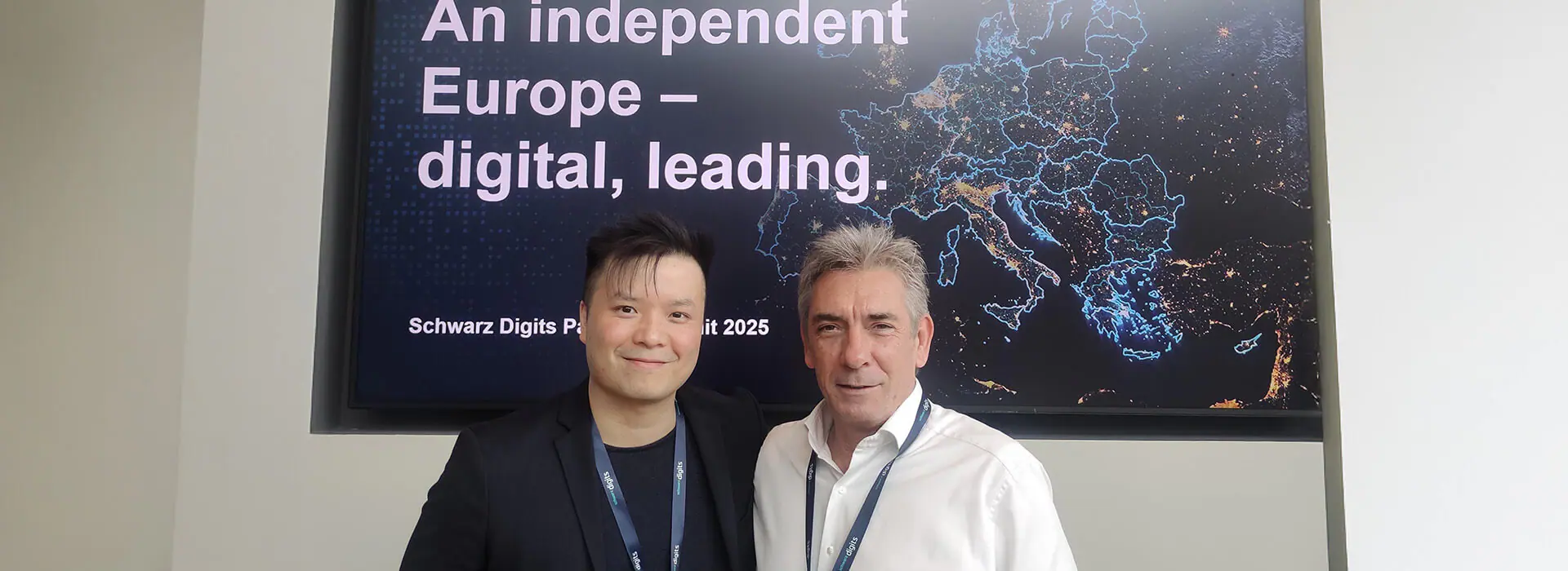 Zwei lächelnde Männer mit Schlüsselbändern stehen vor einem Bildschirm mit einer Europakarte und der Aufschrift "Schwarz Digits Path 2025".