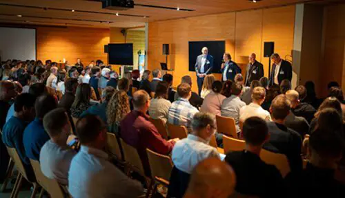 In einem modernen Konferenzraum mit holzgetäfelten Wänden und mehreren Monitoren sitzen die Teilnehmer in Reihen vor fünf Lautsprechern.