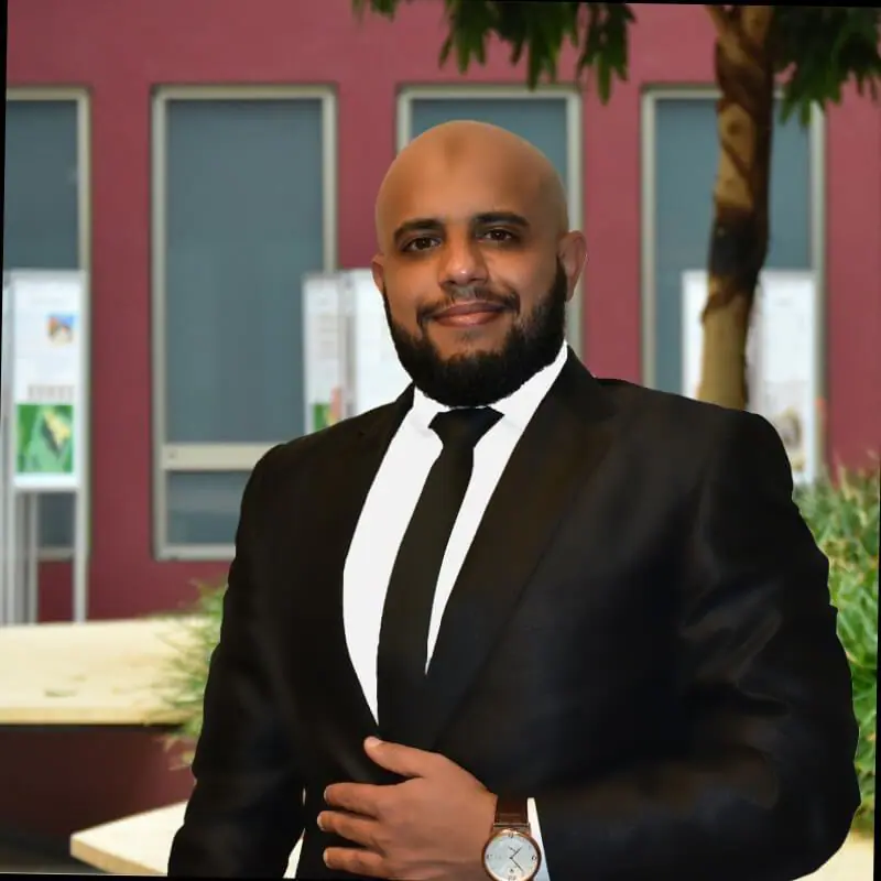 Ein lächelnder Mann mit rasiertem Kopf und Bart in einem schwarzen Anzug steht draußen in der Nähe von Fenstern, Plakaten, einem Baum und Grünpflanzen.