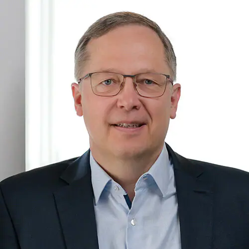 Ein lächelnder Mann mittleren Alters mit kurzen hellbraunen Haaren, Brille, blauem Hemd und dunklem Blazer steht vor einem hellen Hintergrund.