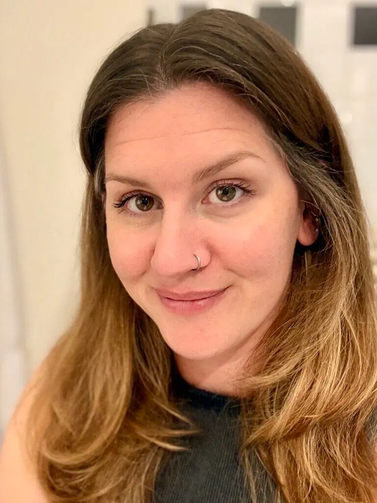 Lächelnde Person mit langen hellbraunen Haaren und einem Nasenring, in einem ärmellosen dunklen Oberteil, in einem Innenraum mit neutralem Hintergrund.