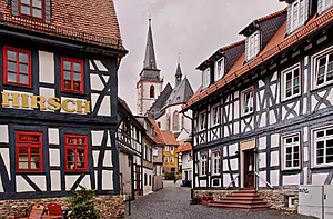 Kopfsteinpflasterstraße mit Fachwerkhäusern, "HIRSCH"-Schild auf der linken Seite und eine Kirche mit hohen Türmen unter einem bedeckten Himmel.