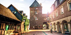 Eine Straße mit Kopfsteinpflaster und Backsteingebäuden führt zu einem mittelalterlichen Torturm mit rot verschlossenen Fenstern, hinter denen das Sonnenlicht scheint.