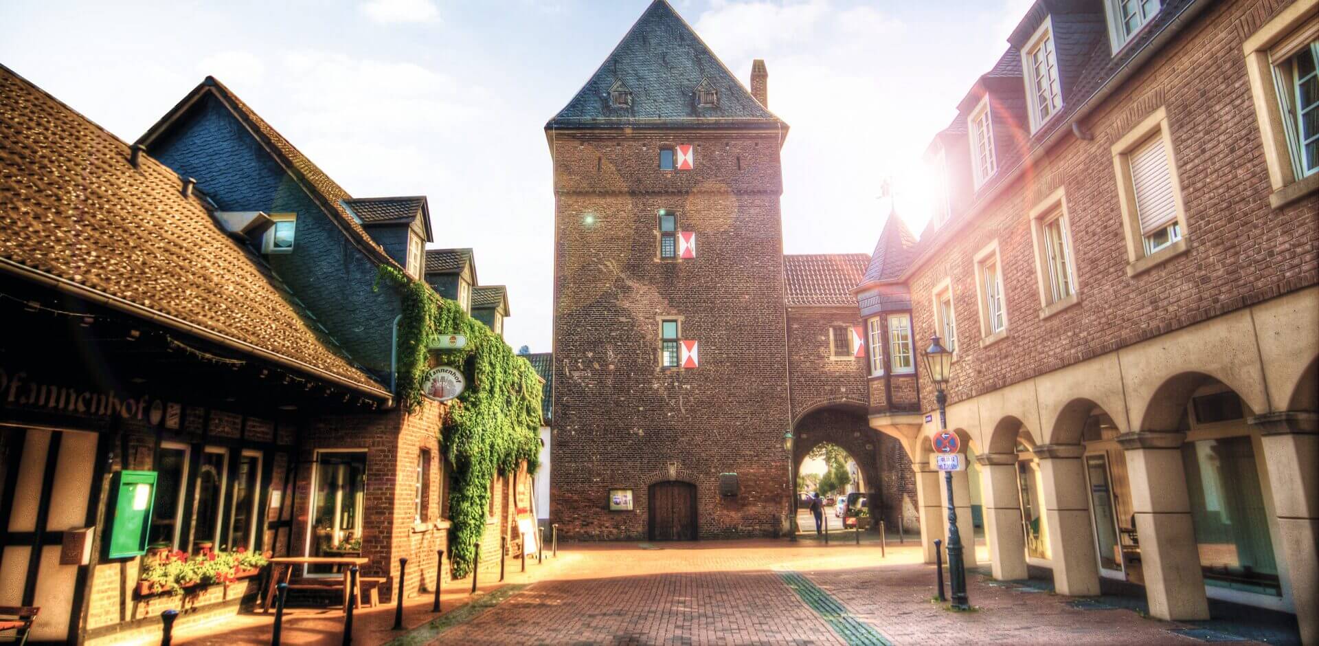 Eine Straße mit Kopfsteinpflaster und Backsteingebäuden führt zu einem mittelalterlichen Torturm mit rot verschlossenen Fenstern, hinter denen das Sonnenlicht scheint.