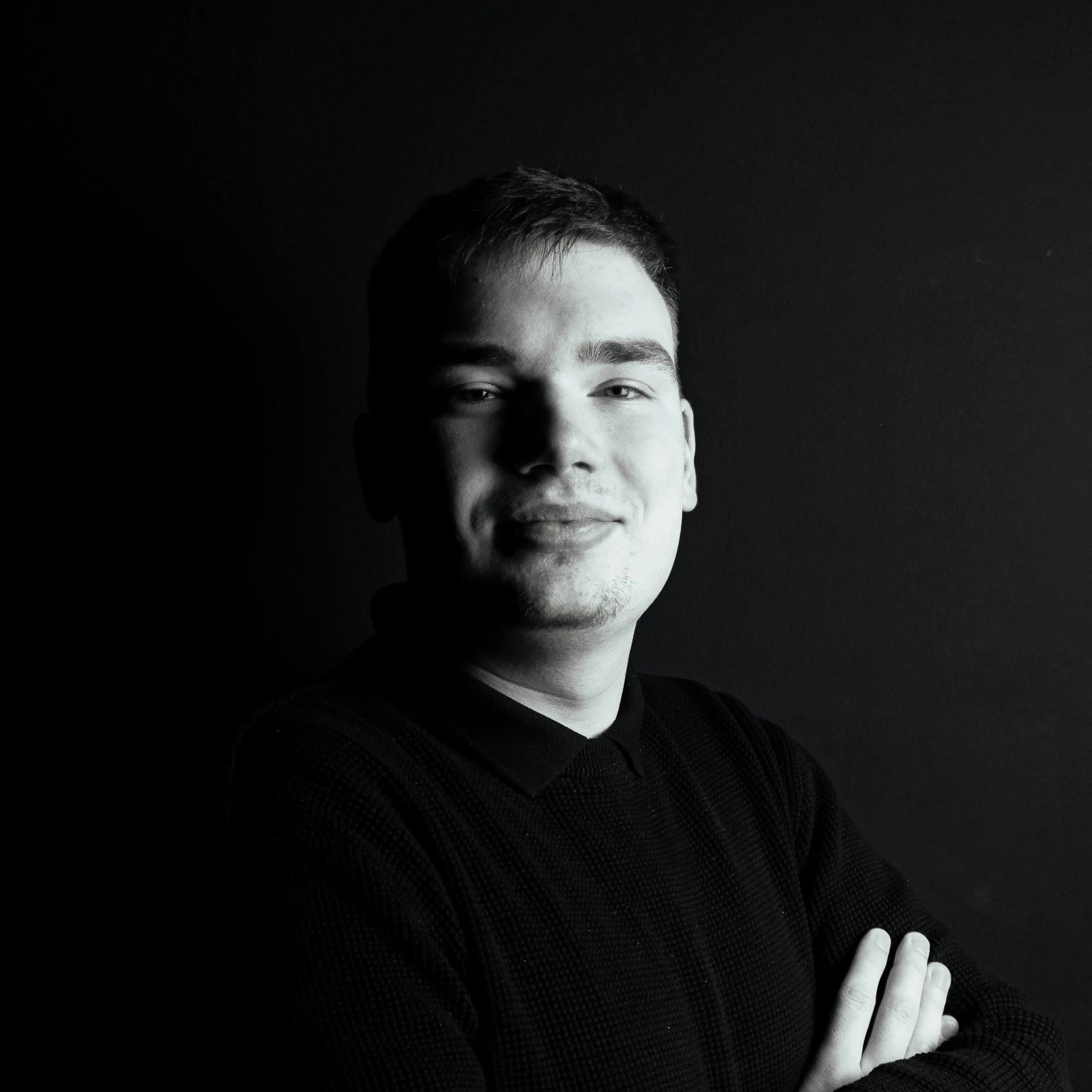 Young man in a dark sweater stands against a black background, arms crossed, one side of his face partially shadowed.