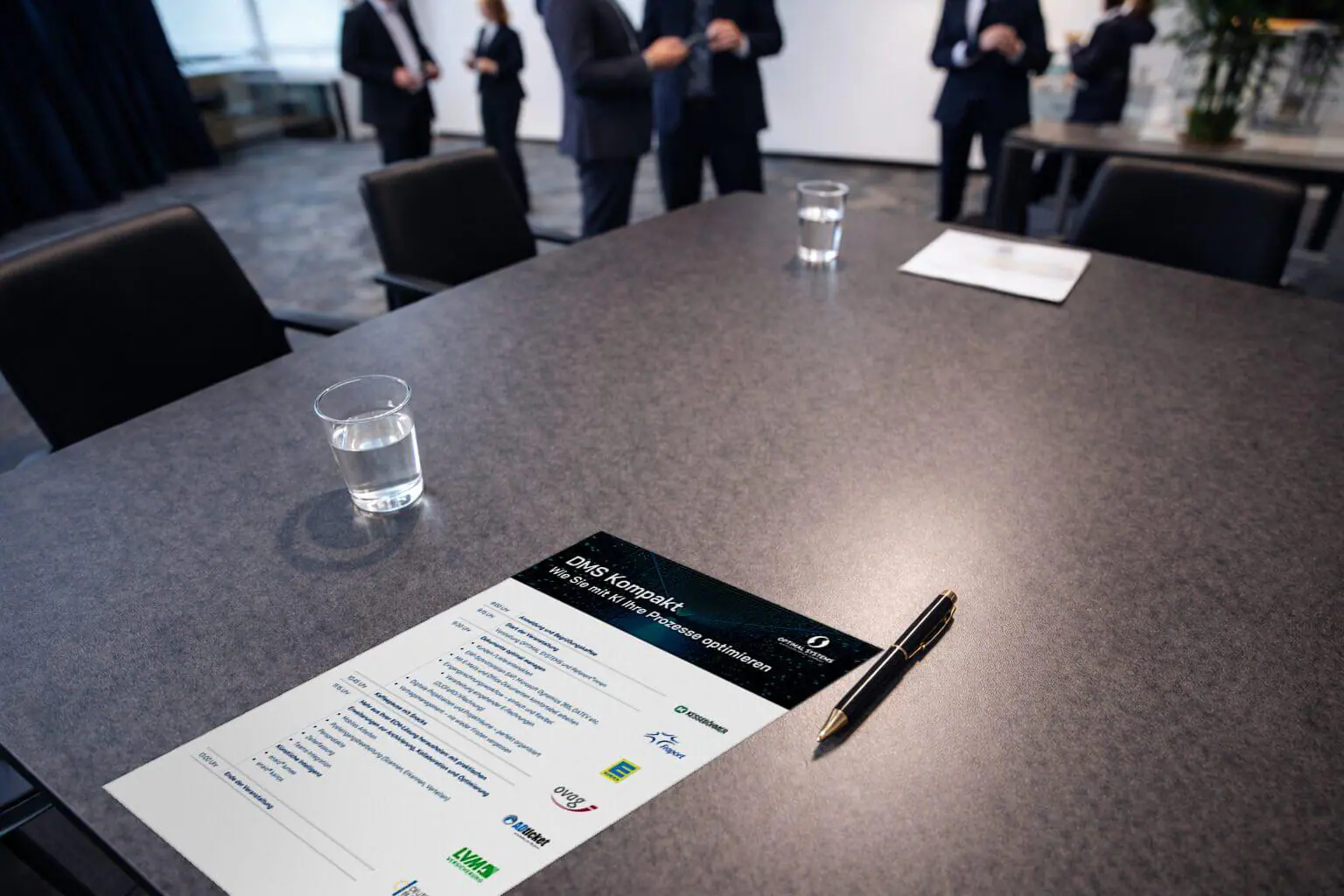 Sechs Personen schauen sich in einem modernen Büro gemeinsam eine Bildschirmpräsentation an, auf dem Tisch stehen Snacks und Getränke, und durch große Fenster fällt Tageslicht.