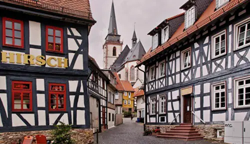 Eine Straße mit Kopfsteinpflaster, flankiert von roten und weißen Fachwerkhäusern, im Hintergrund erhebt sich eine hohe, steile Kirche.