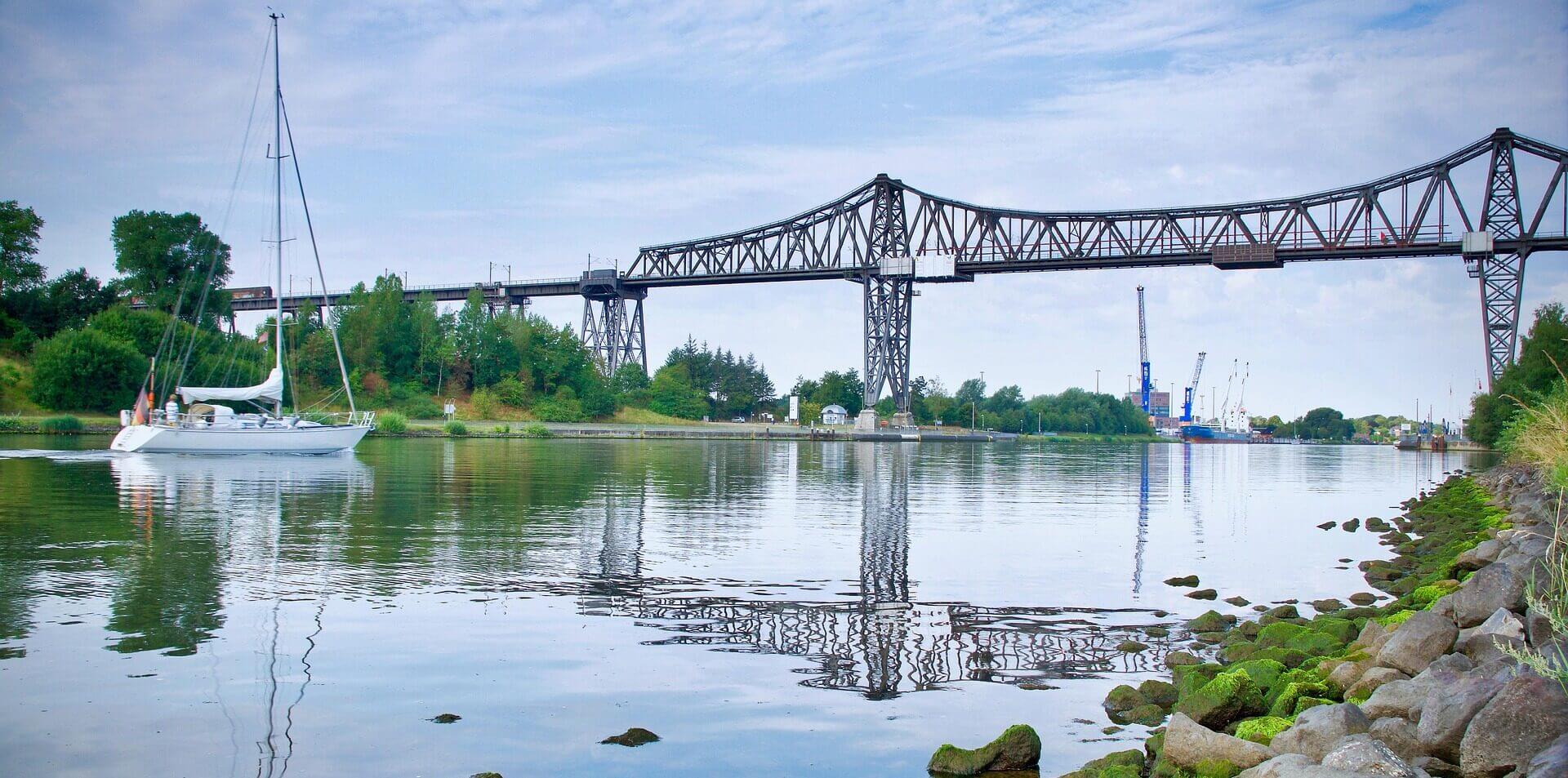 Ein weißes Segelboot auf einem ruhigen Fluss in der Nähe von Felsen, mit einer stählernen Eisenbahnbrücke und Industriekränen im Hintergrund.