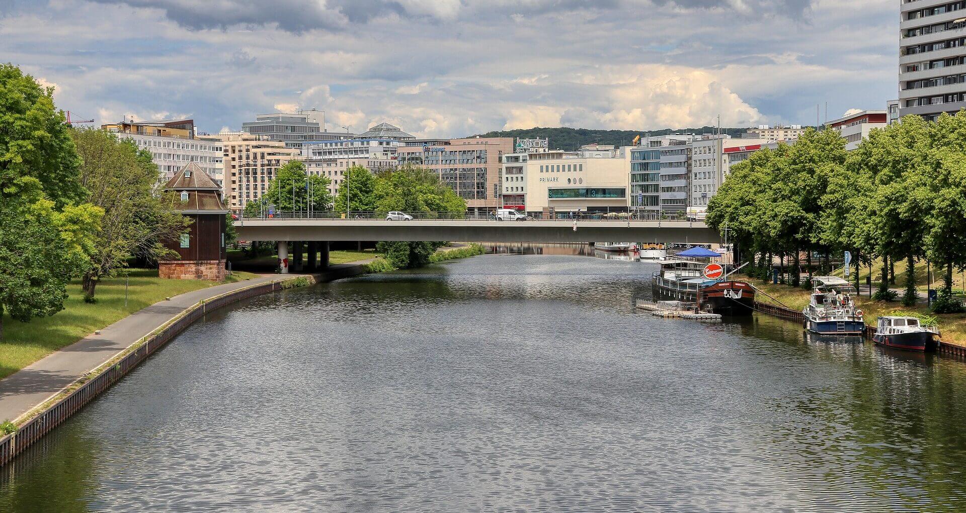 Ein Fluss fließt durch eine Stadt mit modernen Gebäuden, einer Brücke, Booten am rechten Ufer und grünen Bäumen unter einem teilweise bewölkten Himmel.