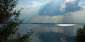 Das Sonnenlicht bricht durch die Wolken über einem ruhigen See, mit Bäumen an den Seiten und hellen Reflexen auf dem Wasser.