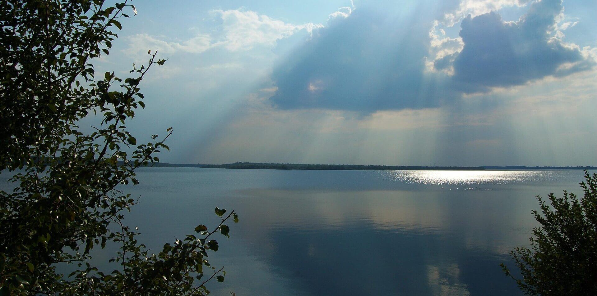 Das Sonnenlicht bricht durch die Wolken über einem ruhigen See, mit Bäumen an den Seiten und hellen Reflexen auf dem Wasser.