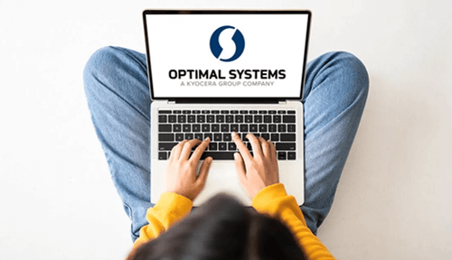 Person in gelbem Pullover sitzt im Schneidersitz und tippt auf einem Laptop mit dem Logo "OPTIMAL SYSTEMS A KYOCERA GROUP COMPANY".